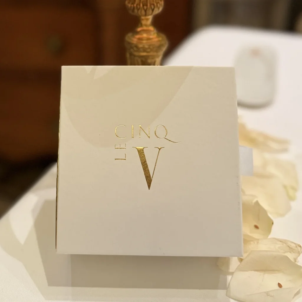 Elegant white gift box with gold Le Cinq logo, presented at the end of dinner on a petal-strewn table.