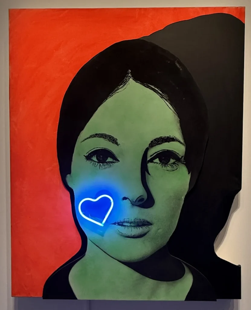 Portrait of a woman with green skin, black hair, and a glowing blue neon heart on her cheek against a vivid red background.