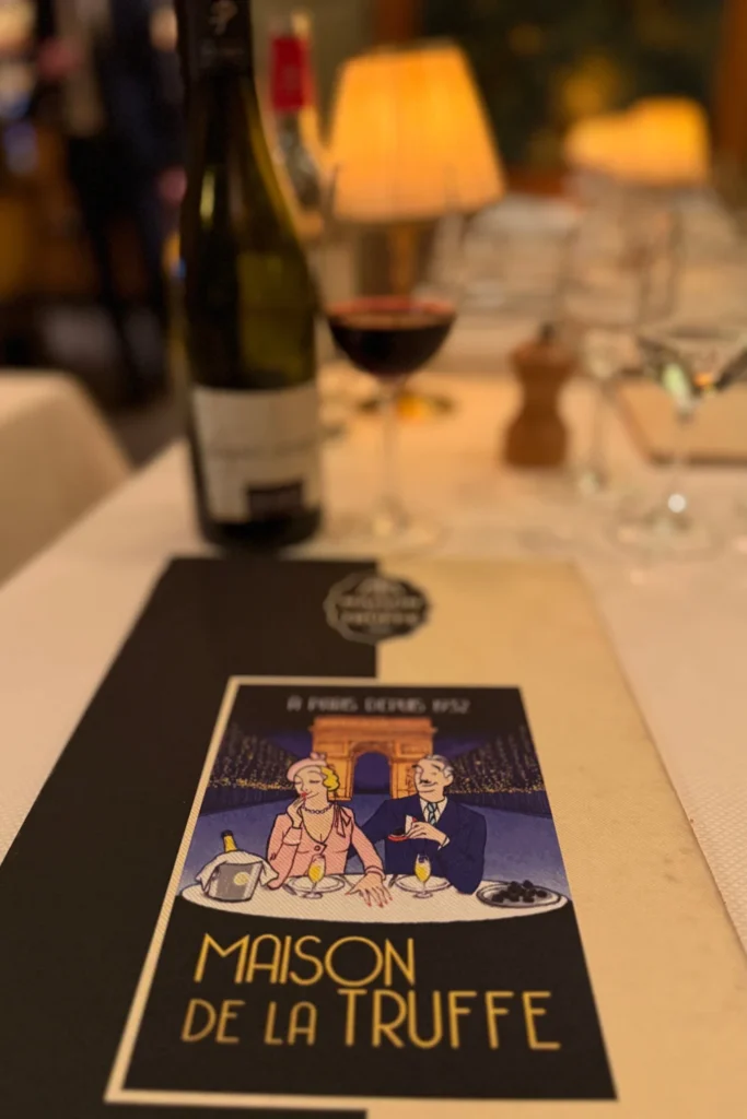 Table at Maison de la Truffe set with house menu and a bottle of Saint-Joseph red wine, softly lit with a table lamp