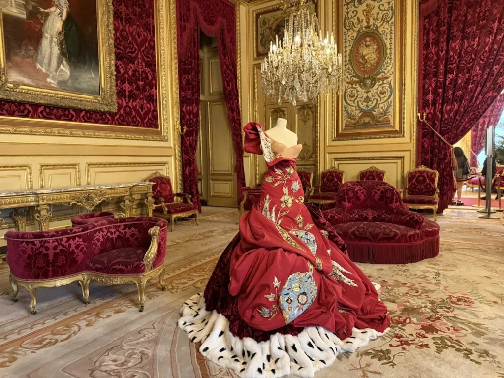 red Dior ballgown with embroidered detailing and ermine trim displayed in an ornate red and gold salon at the Louvre Couture Exhibition