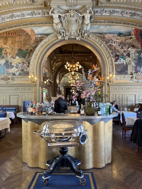 Central bar with gold accents and floral display at Le Train Bleu, framed by painted archways